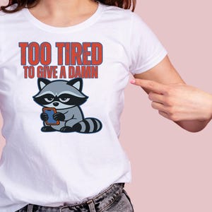 May include: White t-shirt with the text "TOO TIRED TO GIVE A DAMN" in red and blue. The shirt features a cartoon raccoon holding a phone. The raccoon is gray, black, and white.