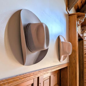May include: Two beige cowboy hats hanging on a wall. The larger hat is on the left and the smaller hat is on the right.