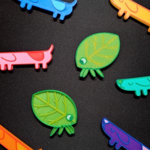 Puede incluir: Una colección de figuritas de colores impresas en 3D, que incluyen un dachshund azul, rosa, naranja, morado y amarillo, y dos criaturas verdes con forma de hoja con ojos.