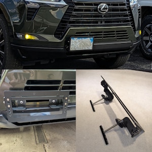 May include: A black metal license plate bracket and mounting hardware. The bracket is designed to attach to the front of a vehicle. The image also shows a close-up of the front of a dark green SUV.