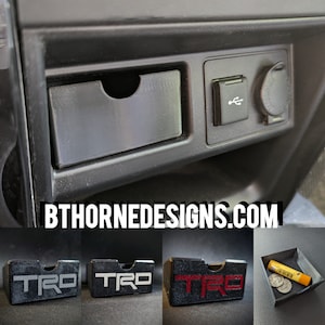 May include: Three black plastic storage containers with the word "TRO" printed on them. The containers are designed to fit in a car's center console. The containers are shown next to a black car console with a USB port and a cigarette lighter.