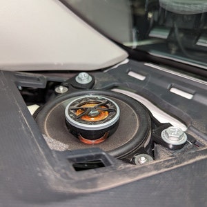 Puede incluir: Primer plano de un altavoz de coche con una rejilla negra y plateada. El altavoz está montado en una carcasa de plástico negro.