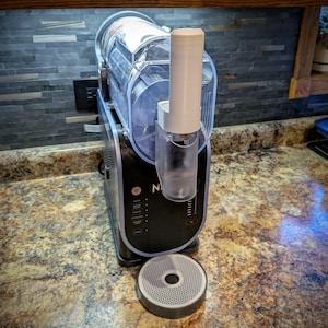 Op de afbeelding: Een zwarte en zilveren elektrische ijsmachine met een doorzichtige plastic container en een witte plastic tuit. De machine staat op een aanrecht met een grijze en witte gepatenteerde basis.