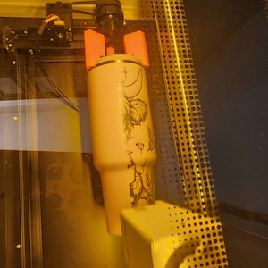 May include: A pink tumbler with a floral design being engraved with a laser cutter. The tumbler is held in place by two pink clamps.