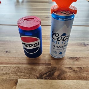 May include: Two beverage cans with custom lids sit on a wooden table. One is a Pepsi can with a red lid, and the other is a Coors Seltzer can with an orange lid. The Coors can is open, and the Pepsi can is closed.