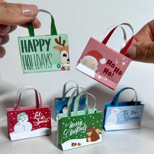 Puede incluir: Cinco pequeñas bolsas de regalo de papel con temas navideños. Las bolsas están decoradas con varios diseños navideños, incluyendo un muñeco de nieve, un reno, Papá Noel, un pingüino y una ardilla. Las bolsas tienen asas y están impresas con mensajes festivos como "Felices Fiestas", "Ho Ho Ho", "Que nieve", "Feliz Navidad" y "Es la temporada".