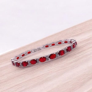Peut inclure: Un bracelet de tennis en argent avec une rangée de pierres précieuses rouges ovales. Le bracelet est posé sur une surface en bois.
