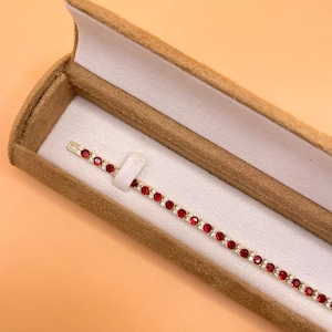 Könnte beinhalten: Ein goldfarbenes Tennisarmband mit abwechselnd roten und klaren Edelsteinen, präsentiert in einer braunen Samt-Schmuckschatulle mit weißem Innenraum. Das Armband liegt diagonal in der Schatulle.