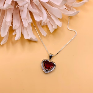 May include: A silver necklace with a red heart pendant. The heart has a silver border with small clear stones.