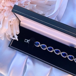 Puede incluir: Una pulsera plateada con piedras preciosas azules en forma de lágrima, cada una bordeada con pequeñas piedras transparentes. La pulsera se exhibe en una caja de joyería negra con interior rosa, sobre un fondo de satén blanco.