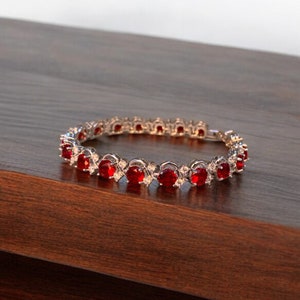 Può includere: Un braccialetto d'argento con una fila di pietre rotonde rosse e trasparenti. Il braccialetto è su una superficie di legno.