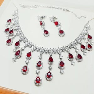 Könnte beinhalten: Ein silberfarbenes Schmuckset mit Halskette und passenden Ohrringen. Die Halskette und die Ohrringe sind mit funkelnden, klaren Steinen und tropfenförmigen roten Edelsteinen verziert. Die Halskette hat eine zarte Kette und eine Reihe von hängenden roten Edelsteinen.