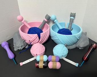 May include: Two yarn bowls, one pink and one blue, shaped like cats with yarn coming out of their backs. Each bowl has a ball of yarn inside and a knitting needle sticking out. There are also four balls of yarn with measuring tape and knitting needles on a gray surface.