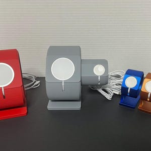 May include: Four different colored stands for charging Apple Watches. The stands are made of a smooth, hard plastic. The stands are in red, gray, blue, and orange.