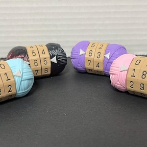May include: Four yarn balls with numbers on them. The yarn balls are blue, pink, purple, and black. Each ball has a number counter that can be turned to count the number of rows of knitting.
