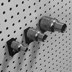 以下が含まれることがあります： 3本の銀色のソケットレンチが、ペグボードの壁に掛けられています。レンチは黒いプラスチック製のホルダーに取り付けられています。ペグボードには、均等に間隔を空けた穴のパターンがあります。