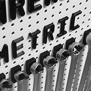 Könnte beinhalten: Schwarzweißfoto einer Lochwand mit dem Wort "METRIC" in schwarzen Buchstaben. Die Lochwand ist mit Schraubenschlüsseln gefüllt, die an Haken hängen.