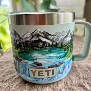 May include: A light green insulated mug with a stainless steel rim and handle. The exterior features a hand-painted landscape of snow-capped mountains, a turquoise lake, and a waterfall. The word YETI is printed in a rectangular white box.