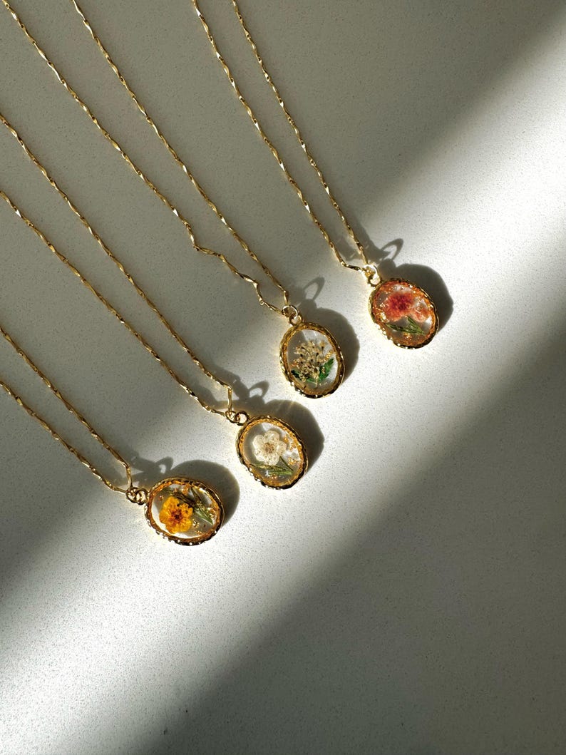 May include: Four gold-toned necklaces with oval pendants, each containing a different pressed flower encased in clear resin. The chains feature a twisted, wavy link design, and the pendants have a textured, metallic gold-colored border.