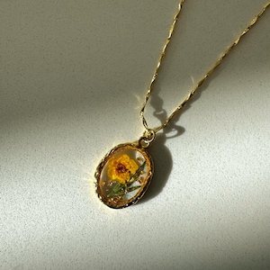 May include: A gold-toned necklace featuring an oval pendant with a textured rim. The pendant contains a pressed yellow flower encased in clear resin, accented with small gold flakes. The chain is a delicate, twisted gold-toned metal link style.