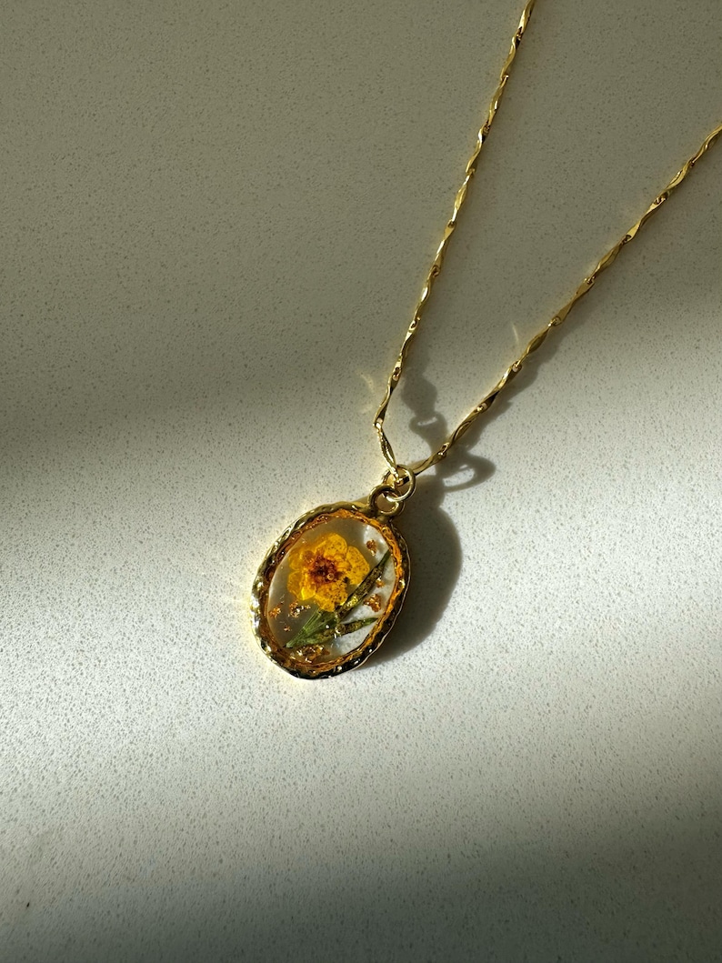 May include: A gold-toned necklace featuring an oval pendant with a textured rim. The pendant contains a pressed yellow flower encased in clear resin, accented with small gold flakes. The chain is a delicate, twisted gold-toned metal link style.