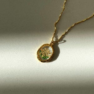 May include: A gold-toned necklace featuring an oval pendant with a textured rim. The pendant contains a clear, resin-like center encasing small, dried white flowers with green stems and leaves. The chain is a delicate, twisted gold-toned metal link.
