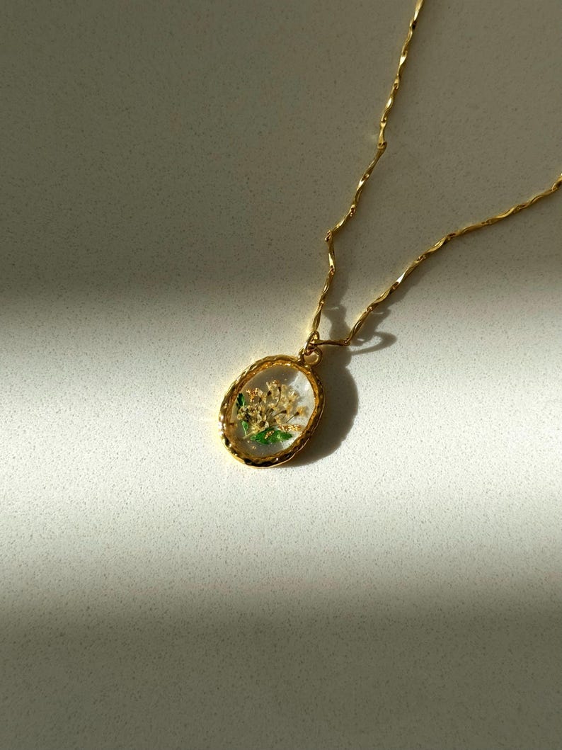 May include: A gold-toned necklace featuring an oval pendant with a textured rim. The pendant contains a clear, resin-like center encasing small, dried white flowers with green stems and leaves. The chain is a delicate, twisted gold-toned metal link.