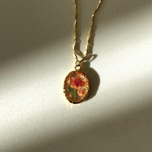 May include: A gold-toned necklace featuring an oval pendant with a scalloped edge. The pendant contains a pressed pink flower with green leaves encased in a clear, resin-like material with gold flakes. The chain is a delicate, twisted gold-tone link.