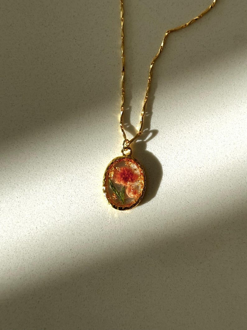 May include: A gold-toned necklace featuring an oval pendant with a scalloped edge. The pendant contains a pressed pink flower with green leaves encased in a clear, resin-like material with gold flakes. The chain is a delicate, twisted gold-tone link.