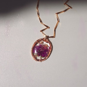 May include: A rose gold necklace featuring an oval pendant with a clear resin center. Inside the resin, a vibrant purple flower is preserved alongside small flecks of gold leaf. The chain consists of thin, angular rose gold metal links.