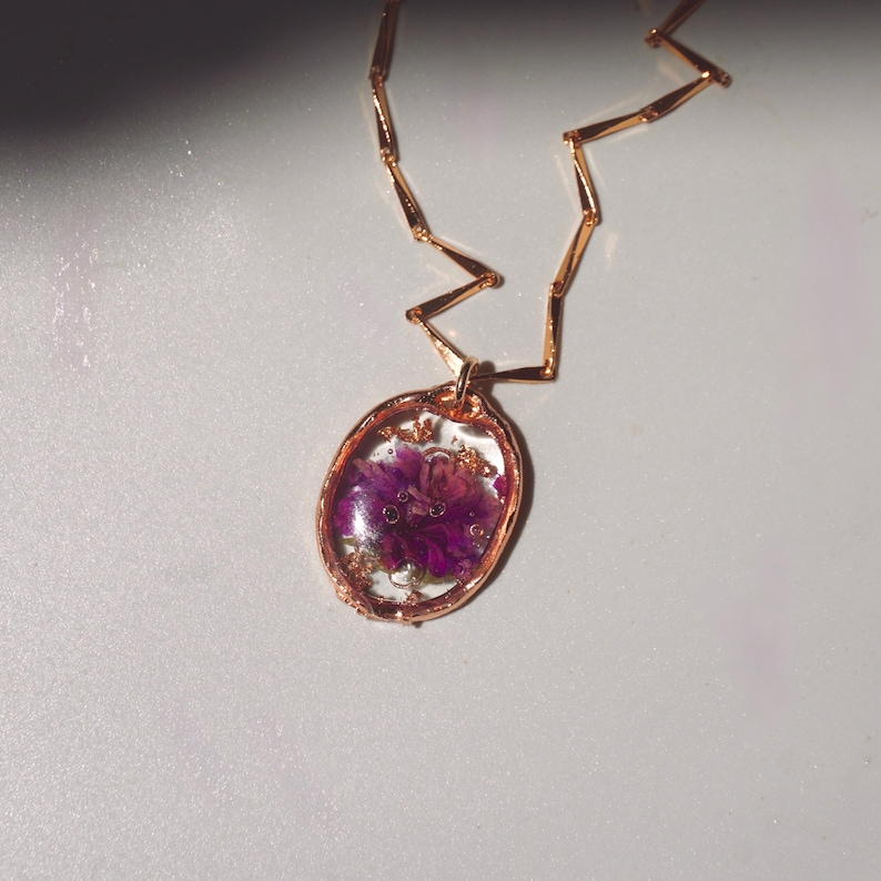 May include: A rose gold necklace featuring an oval pendant with a clear resin center. Inside the resin, a vibrant purple flower is preserved alongside small flecks of gold leaf. The chain consists of thin, angular rose gold metal links.