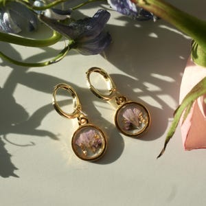 May include: A pair of gold-colored hoop earrings. Each earring features a small, clear, circular pendant containing dried purple flowers and gold flakes. The earrings are displayed on a white surface with floral accents.