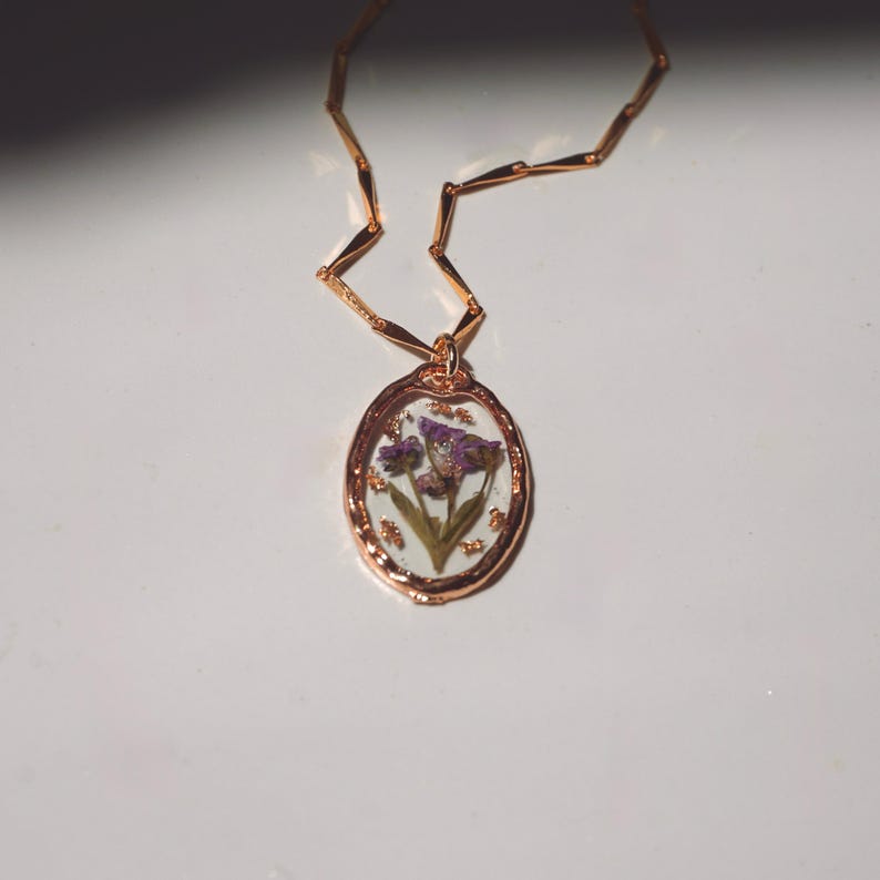 May include: A rose gold necklace featuring an oval pendant with a scalloped edge. The pendant contains a clear resin inlay with pressed purple flowers and small gold-colored flecks. The chain consists of thin, elongated, rectangular links.
