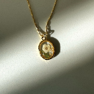 May include: A gold-toned necklace featuring a delicate chain and an oval-shaped pendant. The pendant has a textured gold rim and contains a small, pressed white flower with green stems encased in a clear, resin-like material with gold flakes.