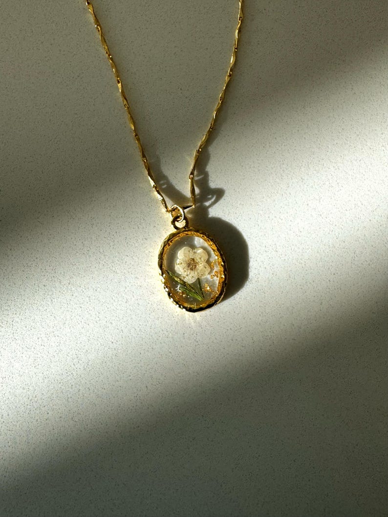 May include: A gold-toned necklace featuring a delicate chain and an oval-shaped pendant. The pendant has a textured gold rim and contains a small, pressed white flower with green stems encased in a clear, resin-like material with gold flakes.