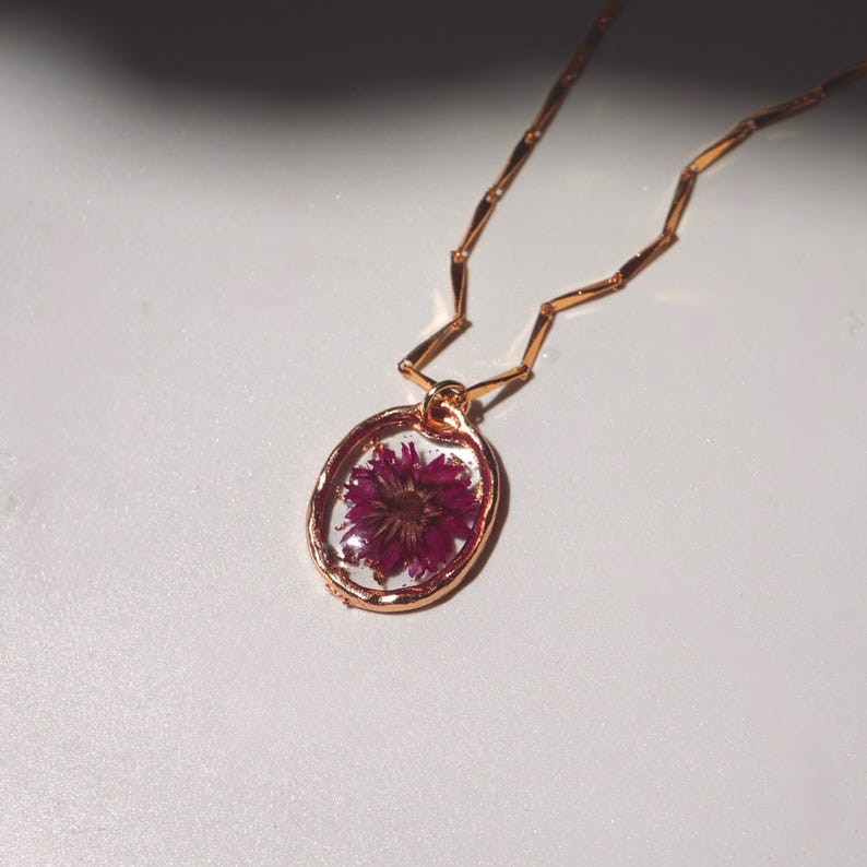 May include: A rose gold necklace featuring an oval pendant with a clear resin center containing a single pressed magenta flower. The pendant hangs from a delicate rose gold chain with small, elongated, rectangular metal links throughout.