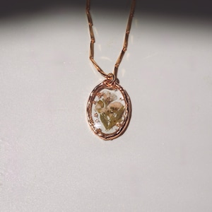 May include: A rose gold necklace featuring an oval pendant with a clear resin center. Inside the resin, small dried white and green flowers are preserved. The pendant is suspended from a delicate rose gold chain with small rectangular links.