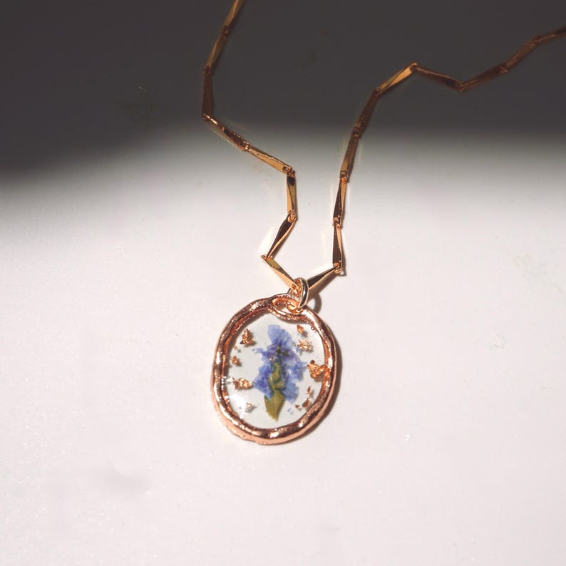 May include: A rose gold necklace featuring an oval pendant with a clear resin center. Inside the resin is a single pressed blue flower surrounded by small gold-colored flakes. The chain consists of thin, flat, rectangular rose gold links.