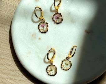 Puede incluir: Cuatro pendientes de aro dorados con un diseño floral. Cada pendiente presenta un pequeño colgante transparente lleno de resina con una flor seca y escamas doradas. Los pendientes se exhiben sobre una superficie de mármol blanco.
