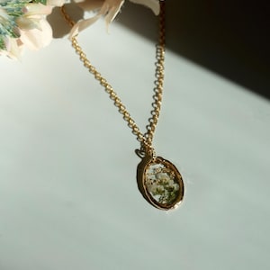 May include: A gold-toned oval pendant necklace featuring a clear resin center that encapsulates small white flowers and green foliage. The pendant hangs from a delicate gold-colored metal chain link, creating a simple and elegant jewelry piece.