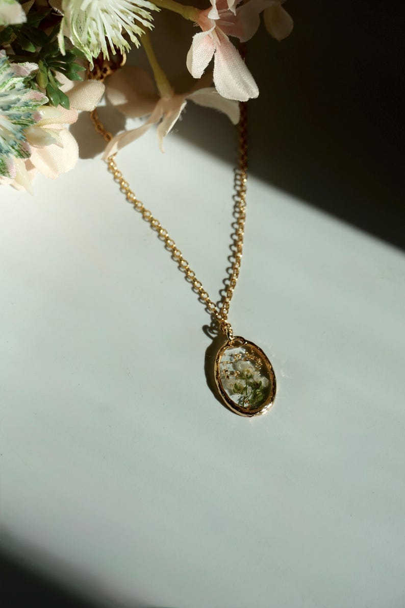May include: A gold-toned oval pendant necklace featuring a clear resin center that encapsulates small white flowers and green foliage. The pendant hangs from a delicate gold-colored metal chain link, creating a simple and elegant jewelry piece.