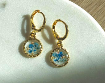 Puede incluir: Pendientes de aro dorados con un pequeño colgante redondo con flores azules prensadas encapsuladas en resina transparente.