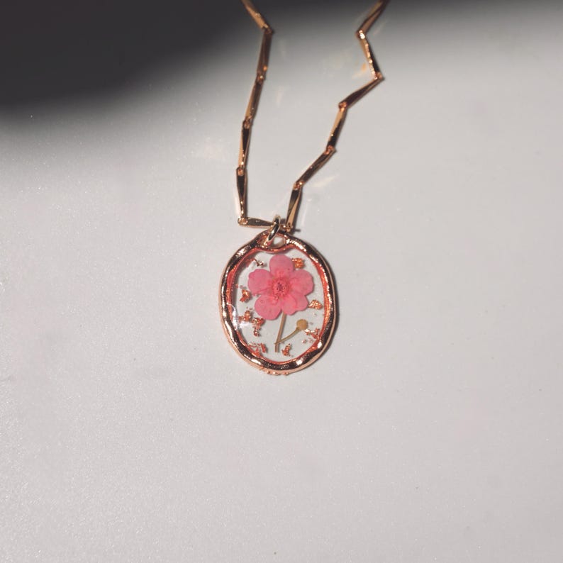 May include: A rose gold necklace featuring an oval pendant with a clear resin center. Inside the resin is a single pink flower with five petals and a thin green stem, accented by small gold flakes. The chain consists of thin, flat, rectangular links.