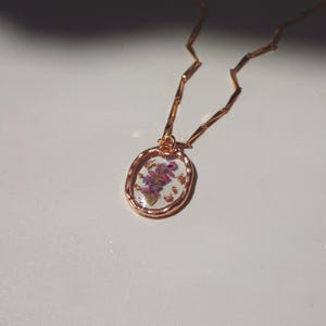May include: A rose gold necklace featuring an oval pendant with a textured metal rim. The pendant contains a clear resin center embedded with small purple dried flowers and tiny gold-colored flakes. The chain consists of thin, flat, angular links.