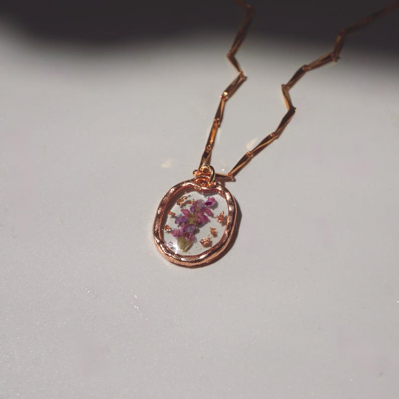 May include: A rose gold necklace featuring an oval pendant with a textured metal rim. The pendant contains a clear resin center embedded with small purple dried flowers and tiny gold-colored flakes. The chain consists of thin, flat, angular links.