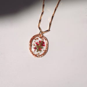 May include: A gold-toned necklace featuring an oval pendant with a textured rim. The pendant contains clear resin embedded with small pink flowers and gold leaf flakes. The chain consists of elongated, flat, rectangular gold-toned links.