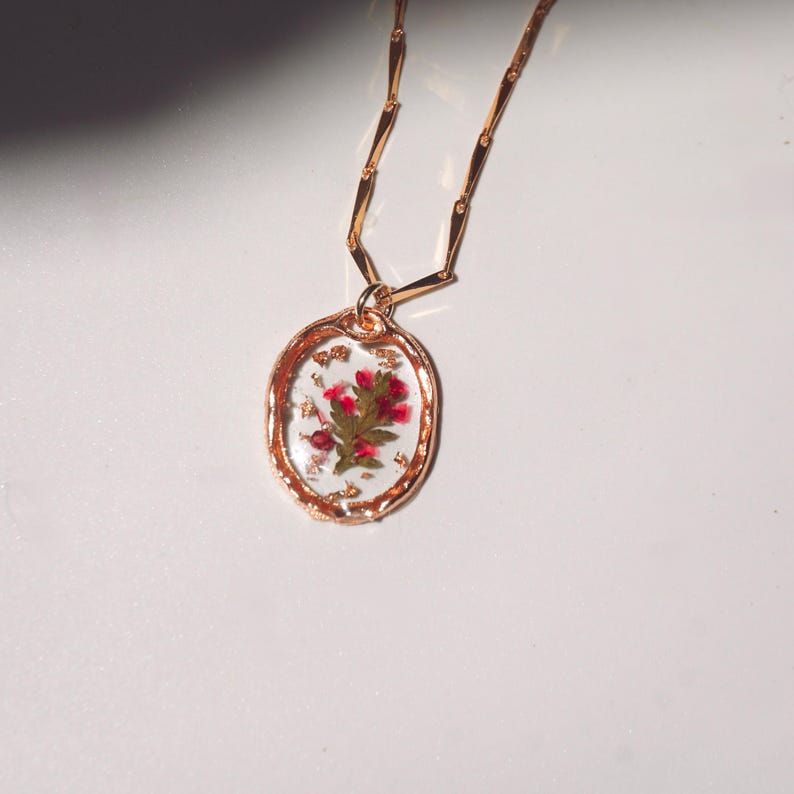 May include: A gold-toned necklace featuring an oval pendant with a textured rim. The pendant contains clear resin embedded with small pink flowers and gold leaf flakes. The chain consists of elongated, flat, rectangular gold-toned links.