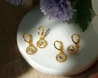 Puede incluir: Cuatro pendientes de aro dorados con pequeños colgantes redondos. Cada colgante presenta flores secas encapsuladas en resina transparente. Los pendientes se exhiben sobre una superficie blanca y circular, con flores moradas en el fondo.