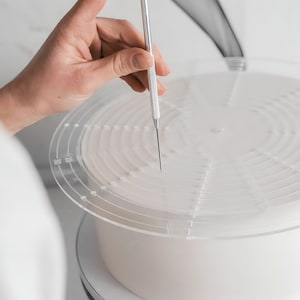 Può includere: Una torta bianca su un supporto rotante per torte con uno strumento di decorazione per torte in acrilico trasparente con uno spillo di metallo. Lo strumento ha cerchi concentrici e punti per marcare la torta.