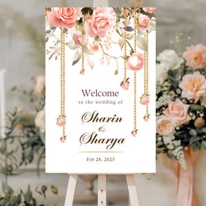 May include: A white wedding welcome sign with pink roses and gold chains. The sign reads "Welcome to the wedding of Sharın & Sharya Feb 28, 2025".